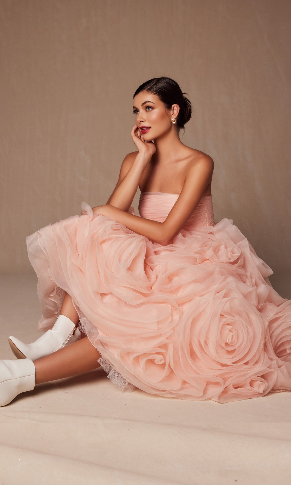 A woman in the Nox Anabel JQ1037 Strapless Peach Prom Ball Gown with ruffled layers and floral details sits on the floor, chin resting on her hand, styled with white ankle boots and pearl earrings.